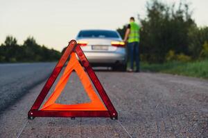 Broken silver luxury car emergency accident. Man driver installing red triangle stop sign on road. Sport automobile turned on blinkers technical problems on the road. Safety procedure when having vehicle broken down photo