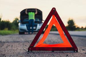 Broken silver luxury car emergency accident. Man driver installing red triangle stop sign on road. Sport automobile turned on blinkers technical problems on the road. Safety procedure when having vehicle broken down photo