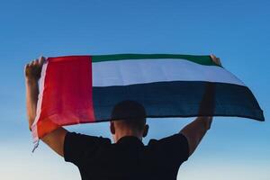 Man holding National Flag Of UAE Waving In The Wind sunset. Flag of United Arab Emirates on Sun Background. Sign of Dubai. Spirit of the Union National Independence Day or Flag day photo