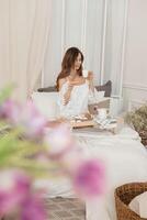 A brown-haired woman with long hair has breakfast in bed with a donut and a cup of coffee. The concept of a cozy home interior photo