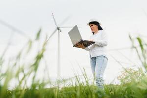 cerca arriba retrato de hembra ingeniero en casco en pie y utilizando ordenador portátil computadora mientras comprobación el trabajo de molino turbina a renovable energía estación. foto