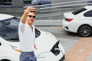 Portrait of young handsome european man, making selfie photo on his smartphone while leaning on his electric car, charging the battery at city power station after buying frsh products in trade mall