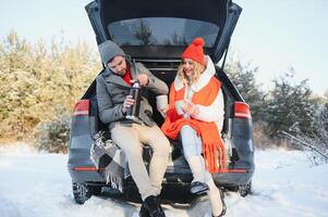 Pareja en amor sentado en coche maletero Bebiendo caliente té en Nevado invierno bosque y charlando personas relajante al aire libre durante la carretera viaje. san valentin día foto