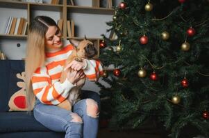 hermosa mujer se sienta en un Clásico sofá con perro. en un antecedentes de un Navidad árbol en un decorado habitación. contento nuevo año foto