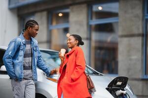 Happy young adult african american man and smiling woman charging electric car. photo