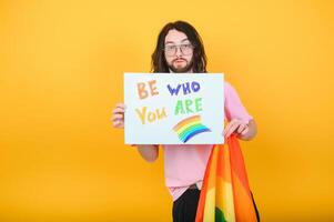 joven queer bisexual sonriente gay hombre con hacer arriba en beige tanque camisa sostener tarjeta firmar con ser quien usted son título texto en arco iris bandera antecedentes estudio retrato. foto