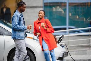 Happy young adult african american man and smiling woman charging electric car. photo