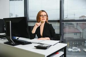 bonito, lindo, lindo, Perfecto mujer sentado a su escritorio en cuero silla en trabajo estación, vistiendo anteojos, Ropa formal, teniendo ordenador portátil y cuaderno en el mesa foto