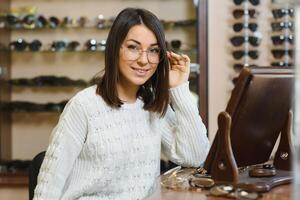 Woman chooses glasses in the store. Brunette in a White sweater buys glasses. Girl on a background of shop windows with different models of glasses photo