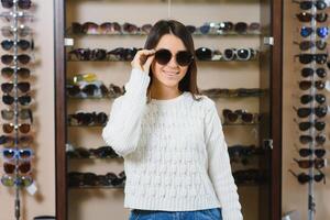 cuales uno es mejor ajuste a mí. retrato de alegre vacilando mujer en óptico almacenar, haciendo decisión, participación elegante Gafas de sol, elegir qué ella debería comprar. genial descuentos para comprando dos pares foto