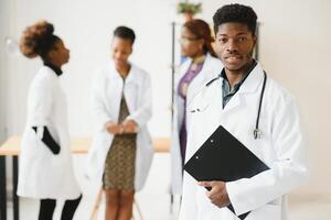 Young African male doctor smiling while standing in a hospital corridor with a diverse group of staff in the background. photo