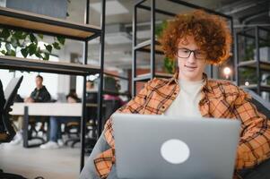 Young happy male freelancer in casual clothes sitting in cafe with laptop and using mobile phone. photo