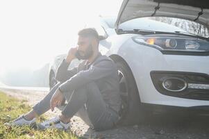 hombre utilizar un Teléfono móvil llamada garaje en frente de el abierto capucha de un roto coche en el la carretera en el bosque. coche Descompostura concepto. foto