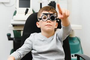 Small serious boy sitting on chair office of vision test. doctor picks up lenses to special glasses. photo