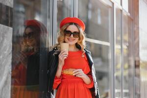 bonito contento mujer con dulce sonrisa en Clásico Gafas de sol en sombrero en casual negro rojo ropa con café camina y sonrisas al aire libre en ciudad. alegre urbano Moda niña en caminar en primavera soleado día. foto