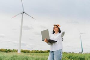 cerca arriba retrato de hembra ingeniero en casco en pie y utilizando ordenador portátil computadora mientras comprobación el trabajo de molino turbina a renovable energía estación. foto