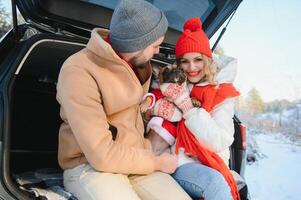 Smiling couple with dog sitting in open SUV car trunk in snowy forest. Enjoying each other in active winter holidays. photo