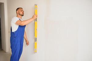 el constructor cuidadosamente corrige el irregularidades de el pared con un paleta. constructor en trabajo ropa en contra un gris pared. foto yesero a trabajar.