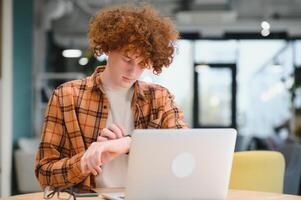 Young happy male freelancer in casual clothes sitting in cafe with laptop and using mobile phone. photo