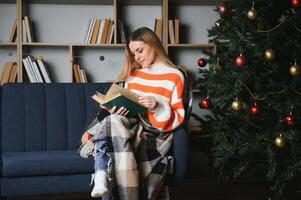 Attractive young lady with dark hair reading interesting book while sitting on grey couch. Blur background of beautiful christmas tree. Cozy atmosphere. photo