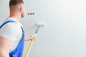 Workman in blue overalls painting the wall with a roller, view from the side photo