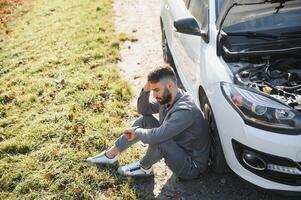 Picture of frustrated man sitting next to broken car with open hood photo