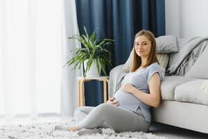 Pregnant woman caressing her belly at home. Young expectant blonde feeling her baby push, sitting on floor, copy space. Pregnancy, rest, life, expectation concept photo