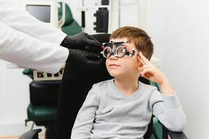 Small serious boy sitting on chair office of vision test. doctor picks up lenses to special glasses. photo