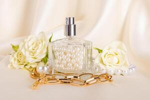 A chic perfume bottle is filled with pearls against the background of beige waves of satin, roses, a gold necklace. Front view. Creative composition. photo