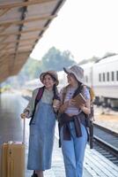 dos joven asiático amigos muchachas con mochilas a ferrocarril estación esperando para tren, dos hermosa mujer caminando a lo largo plataforma a tren estación foto