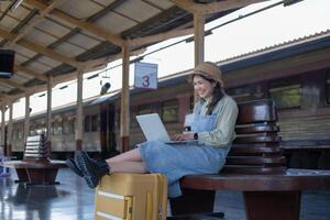 Girl using a laptop while waiting in a train station, Girl on train station with luggage working on laptop computer, laptop in use, sits with a suitcase photo