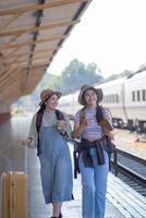 dos joven asiático amigos muchachas con mochilas a ferrocarril estación esperando para tren, dos hermosa mujer caminando a lo largo plataforma a tren estación foto