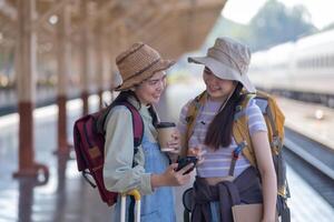 dos joven asiático amigos muchachas con mochilas a ferrocarril estación esperando para tren, dos hermosa mujer caminando a lo largo plataforma a tren estación foto