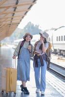 dos joven asiático amigos muchachas con mochilas a ferrocarril estación esperando para tren, dos hermosa mujer caminando a lo largo plataforma a tren estación foto