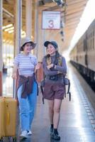 dos joven asiático amigos muchachas con mochilas a ferrocarril estación esperando para tren, dos hermosa mujer caminando a lo largo plataforma a tren estación foto