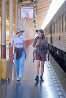 dos joven asiático amigos muchachas con mochilas a ferrocarril estación esperando para tren, dos hermosa mujer caminando a lo largo plataforma a tren estación foto