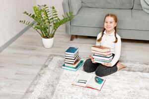 linda pequeño niña sentado en el piso a hogar con un apilar de libros en su manos foto