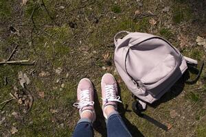 Female legs in jeans and pink sneakers are standing in a ground of forest with backpack. photo