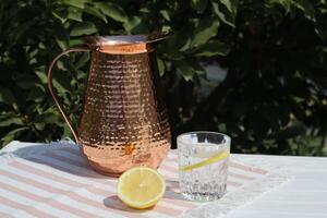 Copper pitcher and water with lemon on the wooden table outdoor. Summer still life with water and pitcher. photo