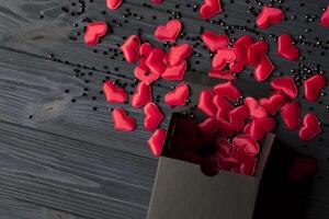 rojo corazones son vertido fuera de un negro regalo caja en un oscuro azul de madera mesa. foto