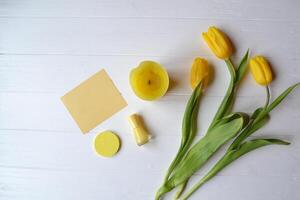 Yellow paper, tulips, candle and female accessories on a desk. Stylish background with place for text. Spring mood card. Yellow still life. photo