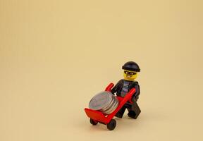 Ukraine, Kyiv - April 6, 2020 Lego robber with a gun takes away coins on a wheelbarrow. Lego man minifigure is carrying money on a trolley. Bank robbery game. Kids toys. Toy figurine man. photo