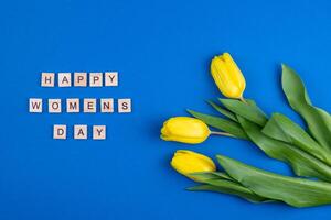 Happy womens day card. Wooden letters. Three yellow spring tulips flowers. Background with flowers tulips close-up. Gift. Blue background. Simple bouquet photo