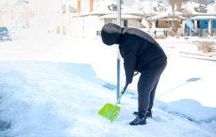 un hombre en un calentar negro chaqueta y pantalones palear nieve en frente de su hogar para preparar el la carretera para conveniencia. foto