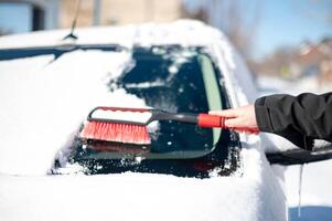 un joven hombre vistiendo negro calentar traje para limpia su coche después un nevada en un soleado, escarchado día. foto