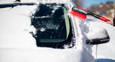 un joven hombre vistiendo negro calentar traje para limpia su coche después un nevada en un soleado, escarchado día. foto