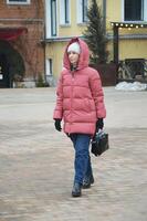 un joven elegante caucásico mujer en un abajo chaqueta, de punto sombrero y pantalones camina a lo largo un ciudad calle en invierno. foto