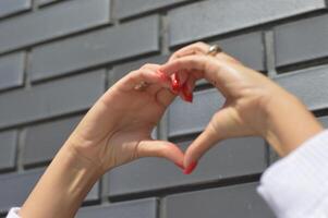 corazón doblada por hembra manos con rojo uñas en contra un gris ladrillo pared. San Valentín día. foto