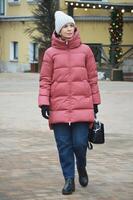 un joven mujer en invierno ropa - un abajo chaqueta, un de punto sombrero, pantalones y con un bolso en su mano es caminando a lo largo un ciudad calle. foto