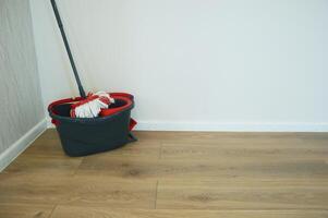 Cleaning mop and black bucket with wringer on wooden floor near white wall. Accessories for cleaning home and office. photo